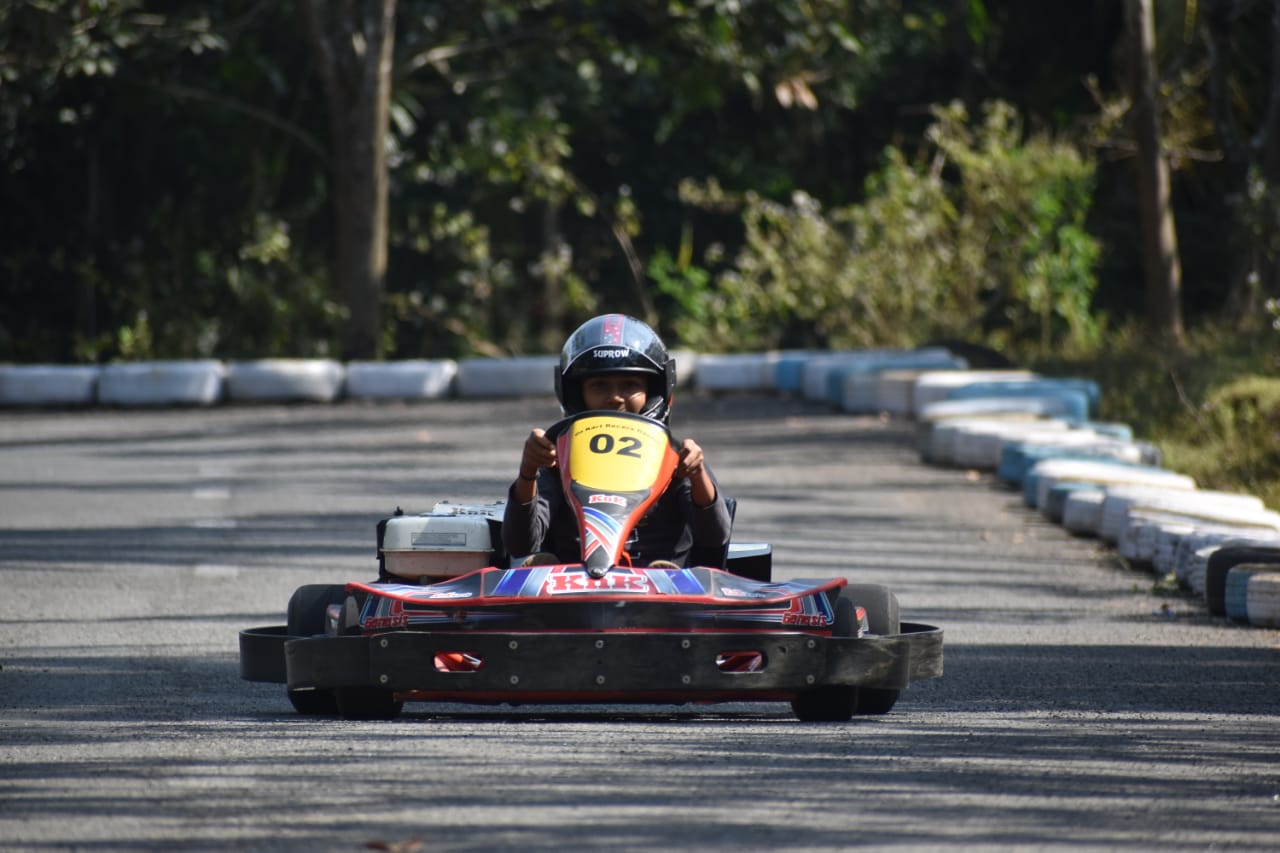 An image of a group of people racing go-karts on a track. The karts are brightly colored and compact, with low-slung bodies and large wheels. The drivers are wearing helmets and racing suits, and are seated low to the ground. They are navigating a twisting track with sharp turns and inclines, surrounded by barriers to keep them on course. The track is brightly lit, and spectators can be seen in the background, cheering on the drivers